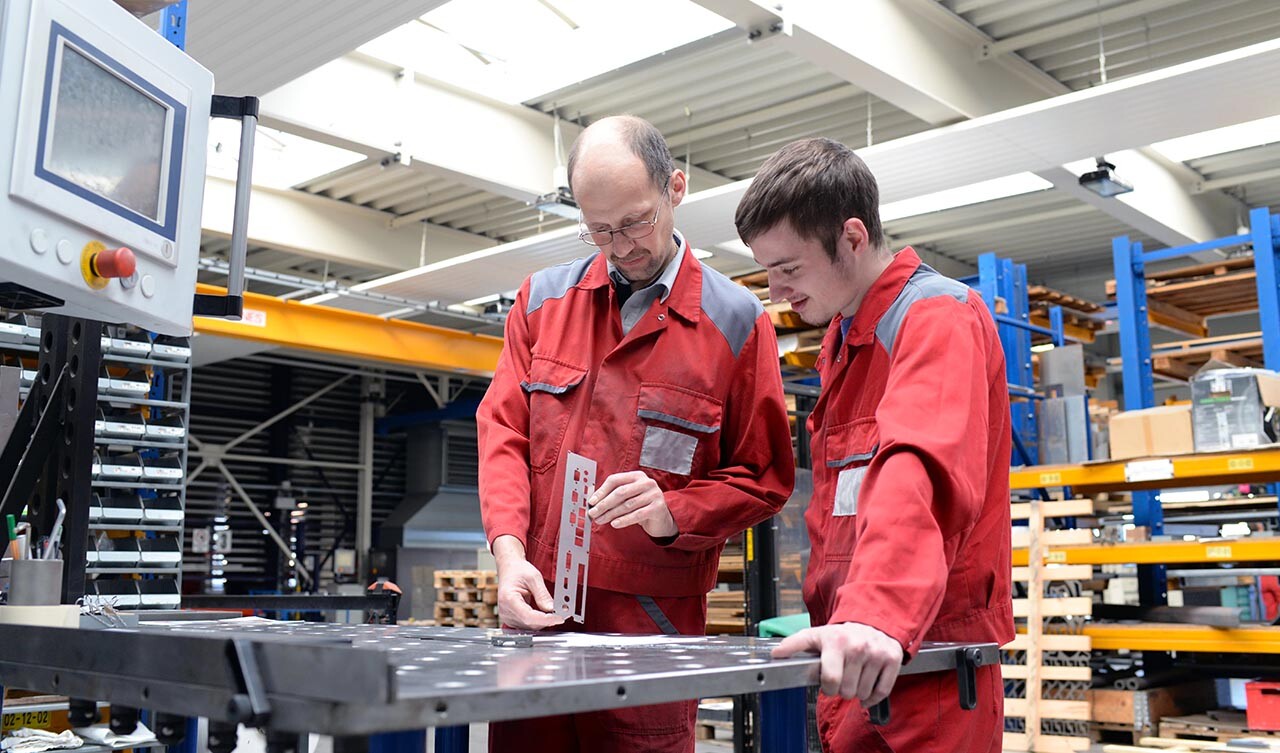 Zwei Mitarbeiter in roten Overalls begutachten ein Stanzprodukt